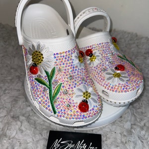 May include: White clogs decorated with embroidered daisies, ladybugs, and rhinestones. The clogs have a white strap and are on a white pedestal. The text "My Style My Way... Be inspired to live the best version of yourself!" is on a black card in the foreground.