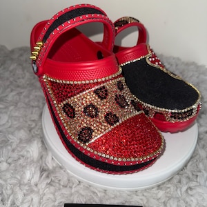 Könnte beinhalten: Rote und schwarze, individuell gestaltete Clogs mit Strasssteinen und einem Leopardenmuster. Die Clogs haben einen schwarzen Riemen und goldene Akzente.