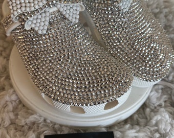 Puede incluir: Zuecos blancos cubiertos de pedrería plateada con lazos blancos en la parte superior. Los zuecos están sobre una superficie blanca con una tarjeta de visita negra que dice "My Style My Way" y "Inspírate para vivir la mejor versión de ti mismo."