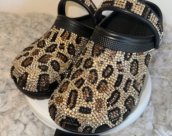 Puede incluir: Zuecos negros y marrones con estampado de leopardo y pedrería. Los zuecos tienen una correa negra y están decorados con pedrería dorada y transparente.