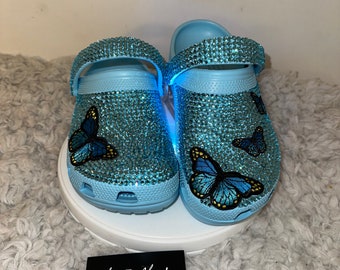 Puede incluir: Zuecos de color azul claro con un diseño de mariposa, cubiertos de pedrería. Los zuecos tienen una correa en la parte superior y están decorados con mariposas azules y amarillas. El texto "My Style My Way" está escrito en una tarjeta negra en la imagen.