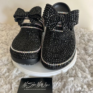 Op de afbeelding: Zwarte clog-sandalen met een strik van strass en een zwart strass-ontwerp. De sandalen liggen op een wit, pluche oppervlak. De tekst "My Style My Way" staat op een zwarte kaart.