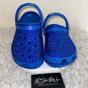 Op de afbeelding: Een paar blauwe klompen bedekt met blauwe strass steentjes. De klompen hebben een bandje aan de achterkant en staan op een wit oppervlak.