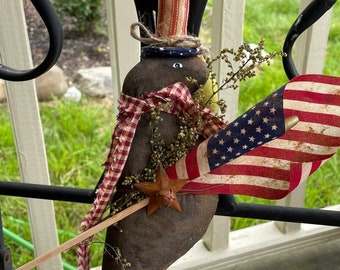 May include: A patriotic-themed folk art bird figurine with a red, white, and blue hat, a red and white checkered scarf, and a small American flag. The bird is holding a wooden stick with a star on it.