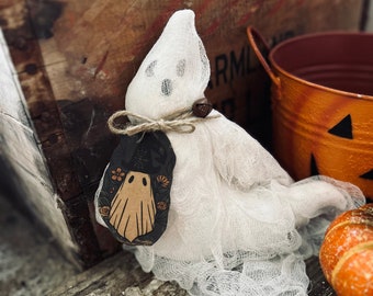 May include: A white cheesecloth ghost figurine featuring a small brown bell tied with twine around its neck. A dark tag hangs from the twine, displaying a graphic illustration of a ghost wearing a hat surrounded by orange and yellow flowers.