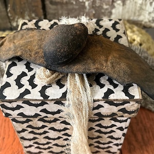 Op de afbeelding: Een vierkante cadeaudoos met een zwart-wit vleermuispatroon. De doos is ingepakt met een bruine vleermuis van stof met een zwarte kop en een stuk jute touw.