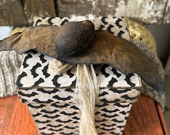 May include: A square gift box with a black and white bat pattern. The box is wrapped with a brown fabric bat with a black head and a piece of burlap twine.