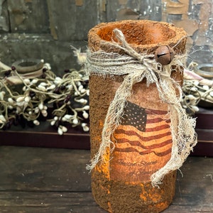 May include: A rustic brown cylinder candle holder with a burlap ribbon and a small bell charm. The holder features a vintage American flag print with the text "United States of America".