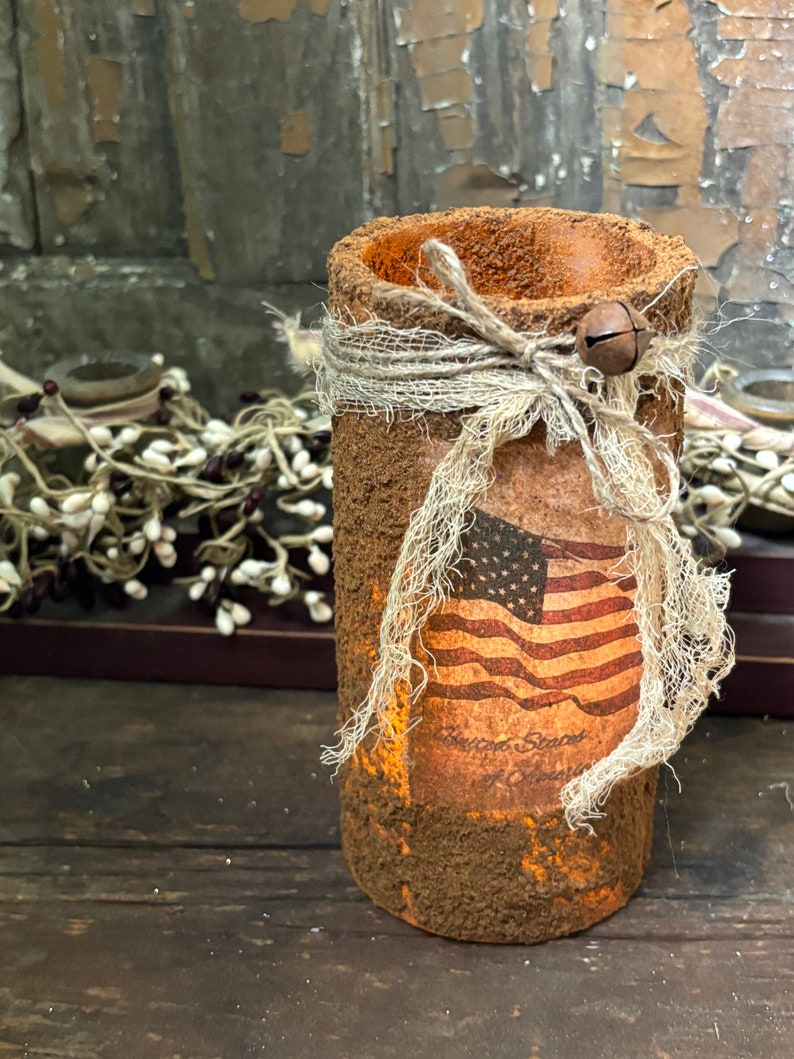 May include: A rustic brown cylinder candle holder with a burlap ribbon and a small bell charm. The holder features a vintage American flag print with the text "United States of America".
