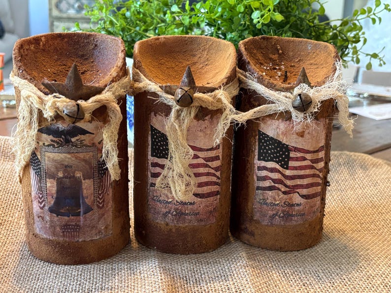 May include: Three rustic brown candle holders with a distressed finish. Each holder features a different patriotic design, including an American flag and a depiction of the Liberty Bell. The holders are decorated with burlap ribbon and a small metal bell.