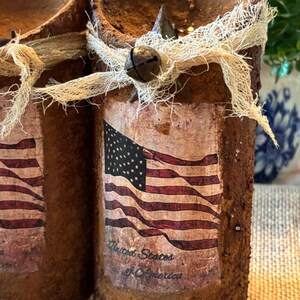 May include: Two rustic brown candles with a distressed finish. Each candle has a burlap ribbon tied around it and a small bell attached. One candle has a vintage American flag design with the text "United States of America".