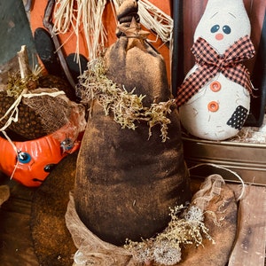 May include: A brown witch's hat with a burlap brim and a brown fabric top. The hat is decorated with dried moss and a small bell. The hat is sitting on a wooden surface with a pumpkin and a ghost doll in the background.