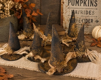 May include: A collection of six miniature witch hats in a dark, distressed black finish with brown accents. Each hat features a pointed crown, a wide brim, and is decorated with frayed tan fabric strips, small dried floral sprigs, and tiny metal bells.