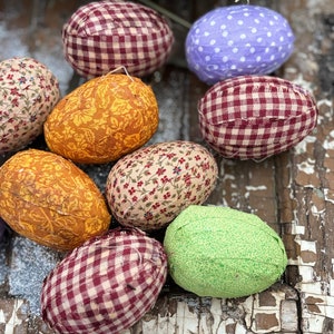 May include: Eight fabric Easter eggs in various patterns, including gingham, floral, and polka dots. The eggs are arranged on a rustic wooden surface.