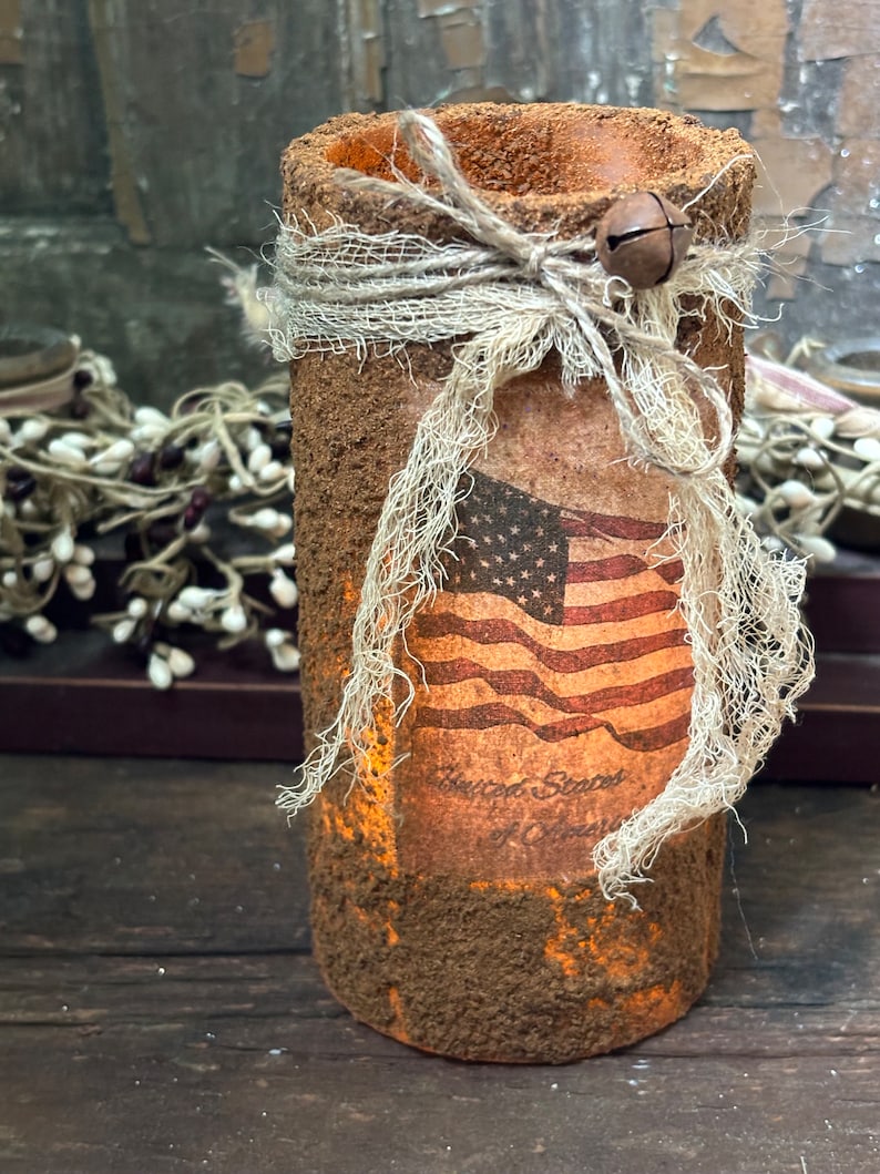 May include: A rustic, brown, cylindrical candle holder with a distressed finish. It is decorated with a burlap ribbon tied with a bow and a small bell. A vintage-style American flag sticker is attached to the front, with the text "United States of America" printed below it.