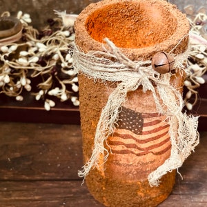 May include: A rustic brown cylinder vase with a burlap ribbon and a small bell charm. The vase has a distressed finish and features a vintage American flag design.