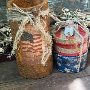 May include: Two patriotic themed containers with red, white, and blue colors. The larger container is a cylinder with a rustic brown finish and a burlap bow. The smaller container is a stack of three round boxes with a red, white, and blue design. The text "United States of America" is visible on the larger container.