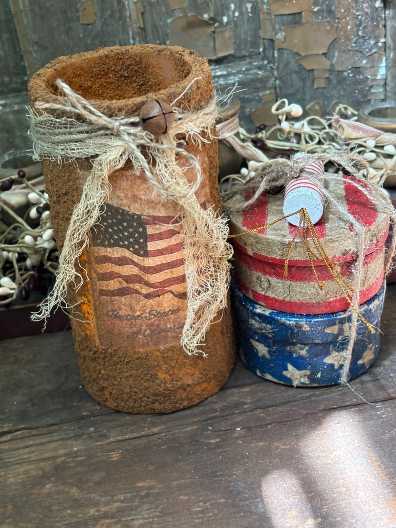 May include: Two patriotic themed containers with red, white, and blue colors. The larger container is a cylinder with a rustic brown finish and a burlap bow. The smaller container is a stack of three round boxes with a red, white, and blue design. The text "United States of America" is visible on the larger container.