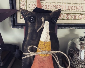 May include: A black fabric cat with a candy corn costume and a witch's hat. The cat has large black eyes and a distressed, vintage look. The candy corn costume is tied with twine.