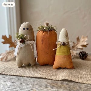 May include: Three fabric ornaments including a beige ghost figure with black eyes and a white scarf, a solid orange pumpkin with a green moss stem, and a candy corn shaped figure with white, yellow, and orange horizontal color bands.