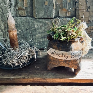 May include: A small black cauldron with a white label that reads "Witchy Brew" is filled with green moss and a small brown twig. A black candle holder with a white candle sits next to the cauldron on a wooden surface.