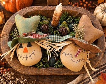 May include: Two stuffed fabric scarecrow heads with painted faces, straw hair, and fabric hats, one green and one brown, resting in a rustic wooden bowl filled with decorative pinecones, greenery, and a single textured pillar candle.
