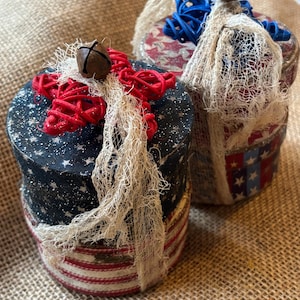 Puede incluir: Dos cajas redondas decorativas con temas patrióticos. Una caja tiene una tapa azul marino con estrellas blancas y un lazo rojo. La otra tiene un diseño de estrellas y rayas rojas, blancas y azules. Ambas tienen una campana en la parte superior.