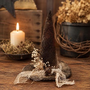 May include: A dark brown, cone-shaped witch hat decoration featuring a textured, weathered finish. The hat is adorned with a small cluster of dried white flowers, a piece of frayed, light-colored mesh fabric, and a single rusted metal jingle bell.