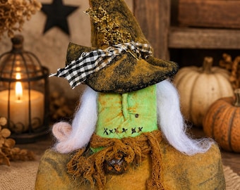 May include: A fabric doll featuring a pointed hat with a black and white checkered ribbon, white yarn hair, and a green torso with stitched X details. The doll has a brown, textured base, a frayed brown fabric sash, and a small metal bell attached.