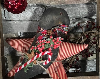 Op de afbeelding: Een rustieke kerststerdecoratie met rood-wit gestreepte stoffen punten en een zwart midden. Het is versierd met een geruite strik, bessen en een zuurstok. Twee rode glazen ballen hangen erboven.