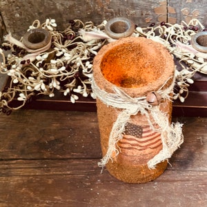 May include: A rustic brown candle holder with a burlap ribbon and a small American flag design. The holder is made of a rough, textured material and has a wide opening at the top.