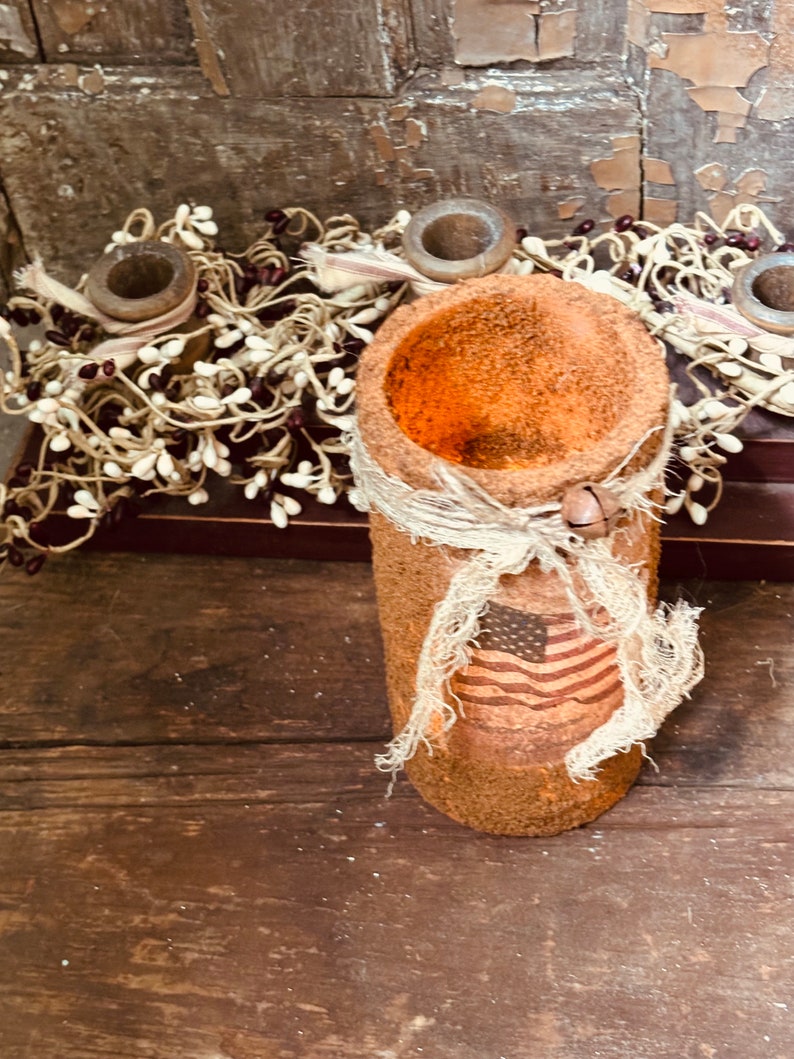 May include: A rustic brown candle holder with a burlap ribbon and a small American flag design. The holder is made of a rough, textured material and has a wide opening at the top.