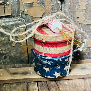 May include: Three stacked round boxes decorated with red, white, and blue patriotic colors. The top box has a red, white, and blue striped design, the middle box has a white and red striped design, and the bottom box has a blue and white star design. The boxes are tied together with twine and topped with two red and white striped paper tubes.