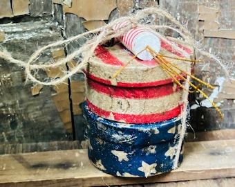 Puede incluir: Tres cajas redondas apiladas decoradas con colores patrióticos rojo, blanco y azul. La caja superior tiene un diseño a rayas rojo, blanco y azul, la caja del medio tiene un diseño a rayas blanco y rojo, y la caja inferior tiene un diseño de estrellas azul y blanco. Las cajas están atadas con un cordel y rematadas con dos tubos de papel a rayas rojo y blanco.