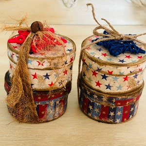 May include: Two stacked round boxes with a patriotic theme. The boxes are decorated with red, white, and blue stars and stripes. The top of each box is adorned with a burlap bow and a star-shaped embellishment. One box has a red star and the other has a blue star.