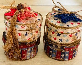 Puede incluir: Dos cajas redondas apiladas con un tema patriótico. Las cajas están decoradas con estrellas y rayas rojas, blancas y azules. La parte superior de cada caja está adornada con un lazo de arpillera y un adorno en forma de estrella. Una caja tiene una estrella roja y la otra una estrella azul.