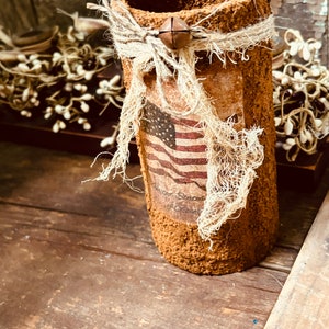 May include: A brown, textured, cylindrical vase with a burlap bow and a bell charm. The vase is decorated with a vintage American flag print and the text "God Bless America".