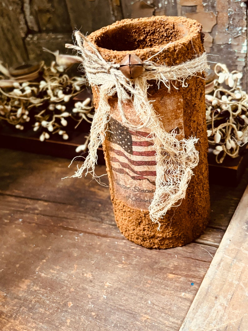 May include: A brown, textured, cylindrical vase with a burlap bow and a bell charm. The vase is decorated with a vintage American flag print and the text "God Bless America".