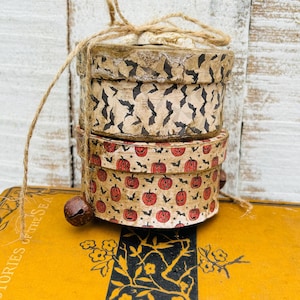 May include: Two stacked round cardboard boxes tied with natural jute twine. The top box features a black bat pattern on a beige background. The bottom box displays a pattern of orange jack-o'-lanterns and small black bats. A dark metal bell is attached.