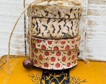 May include: Two stacked round cardboard boxes tied with natural jute twine. The top box features a black bat pattern on a beige background. The bottom box displays a pattern of orange jack-o'-lanterns and small black bats. A dark metal bell is attached.