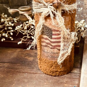 May include: A brown cylindrical vase with a burlap bow and a bell charm. The vase has a distressed finish and features a printed image of the American flag with the text "United States of America".