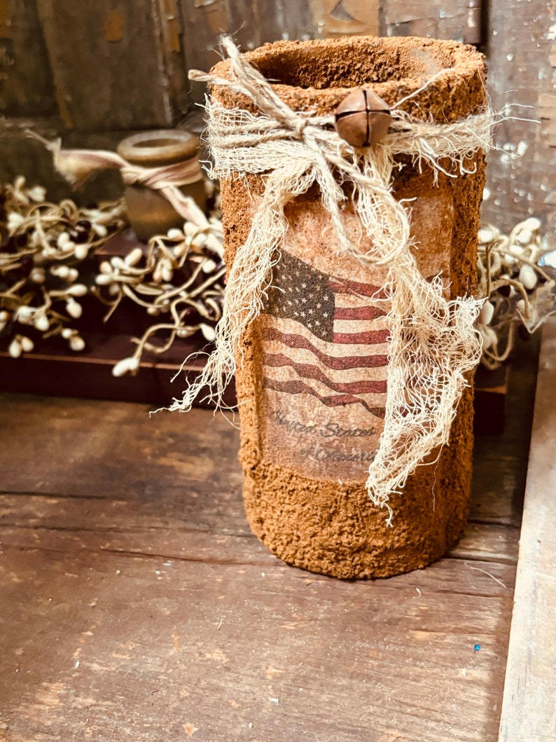 May include: A brown cylindrical vase with a burlap bow and a bell charm. The vase has a distressed finish and features a printed image of the American flag with the text "United States of America".