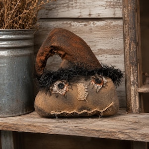 May include: A brown fabric scarecrow figure with a pointed hat, black button eyes, a hessian nose, and a stitched mouth. The figure features frayed black trim around the hat brim and a textured, weathered appearance with visible fabric seams.