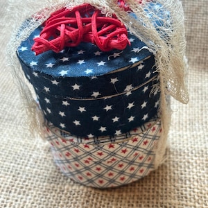 May include: Stacked, round decorative boxes with patriotic themes. The top box is navy blue with white stars, the middle box is navy blue with white stars, and the bottom box is red and white patterned. A red raffia bow and cream-colored netting adorn the top.