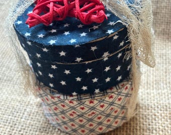 Puede incluir: Cajas decorativas redondas apiladas con temas patrióticos. La caja superior es azul marino con estrellas blancas, la caja del medio es azul marino con estrellas blancas y la caja inferior tiene un estampado rojo y blanco. Un lazo de rafia rojo y una red color crema adornan la parte superior.
