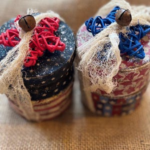 May include: Two round decorative boxes with patriotic designs. One box features a red, white, and blue American flag pattern, while the other has a red and blue star pattern. Each box is adorned with a woven star, a bell, and a cream-colored mesh.