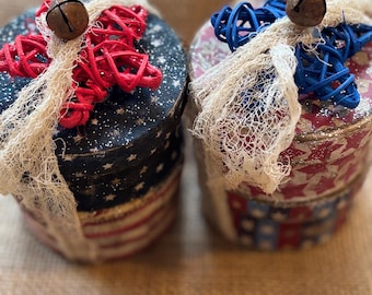 Puede incluir: Dos cajas redondas decorativas con diseños patrióticos. Una caja presenta un patrón de bandera estadounidense roja, blanca y azul, mientras que la otra tiene un patrón de estrellas rojas y azules. Cada caja está adornada con una estrella tejida, una campana y una malla color crema.