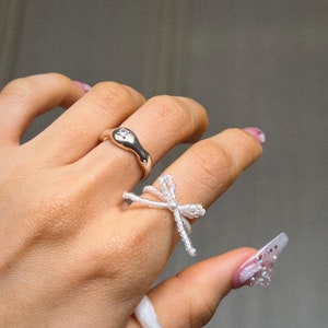 May include: A hand wearing three rings. The ring on the index finger is a silver ring with a small stone. The ring on the middle finger is a white beaded ring shaped like a bow. The ring on the little finger is a white beaded ring.