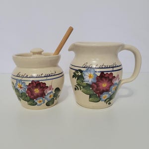 May include: A cream-colored ceramic sugar bowl with a lid and a wooden spoon, paired with a matching creamer pitcher. Both pieces feature hand-painted floral designs in red, blue, and green, with the text Be of a sweet spirit and Perfect strength.