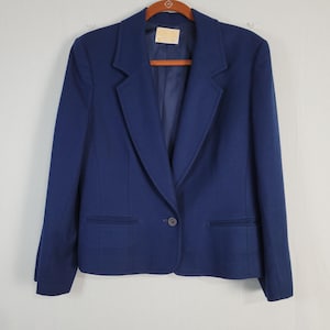 May include: A navy blue blazer with a notched lapel, two front pockets, and a single-button closure. The blazer is hanging on a wooden hanger. The garment has a tailored fit and a classic design.